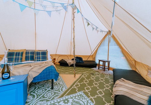 A colourful interior with 4 patterned rugs to offer underfoot comfort and warmth. Cotton Canvas tent sides reveal the full height inside the tent with pale blue bunting dressing the interior tent poles. Four floor mattresses can be seen on either side of the tent next with their own designated and ample space around them. 
A dressed double bed with additional blankets and pillows brings extra home comfort to this camping accommodation.