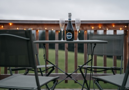 Metal framed and outdoor fabric seating area on the wooden veranda of the wood fired hot tub. Four chairs and one square table make for an ideal spot to sit and watch the clouds roll in as they meet the rolling hills surrounding them, making for a cosy, winter atmosphere. Two champagne flutes and a champagne bottle sit in the centre of the table.