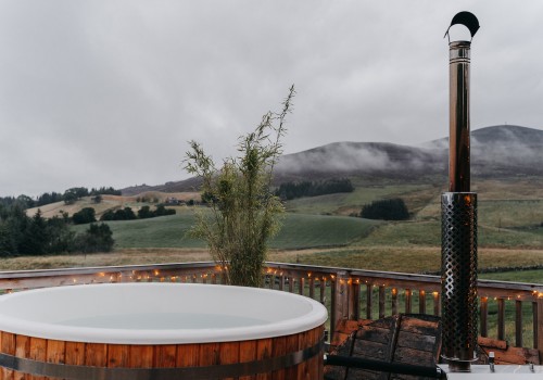 A fully wooden hot tub with its own raised veranda, lit up with cosy lights. Light steam coming off the hot water and low level clouds hitting the rolling hills in the background make for a moody, yet cosy atmosphere. A metal frame table and chairs for two sit in the corner edge of the veranda with a bottle of champagne and two glasses.