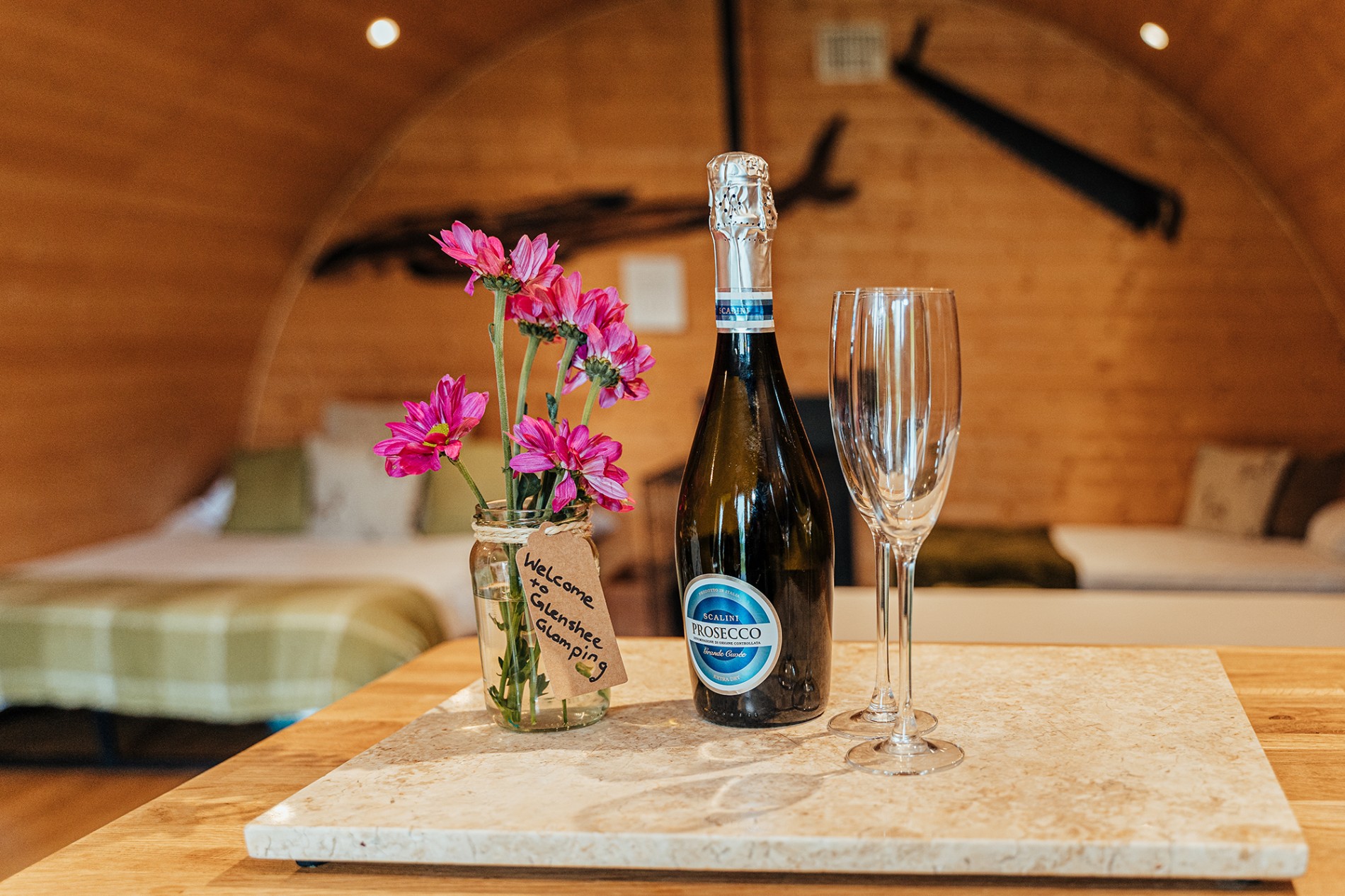 A picture of Welcome champagne, two glassed and a jar of freshly picked flowers from Glenshee Glamping fields sit on the kitchenette bench as you enter the Mulberry accommodation cabin