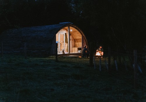 An evening photograph of The Mulberry Cabin Accommodation lit up with warm lights glowing from the wooden cabin. Two guests are sitting outside the glamping cabin around the open fire pit.